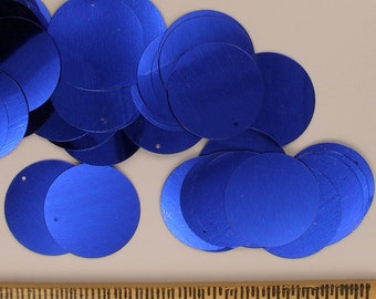 May include: A collection of bright blue, circular sequins with a metallic sheen. Each sequin has a small hole near the edge. The sequins are scattered across a neutral surface, with a ruler visible at the bottom.