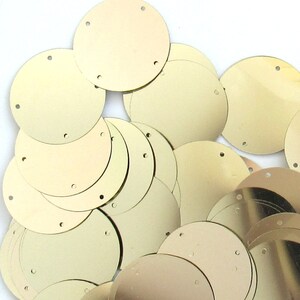 May include: A collection of gold-colored, round sequins with three small holes evenly spaced around the edge. The sequins are scattered across a white surface, creating a textured, reflective appearance. Ideal for crafting and embellishments.