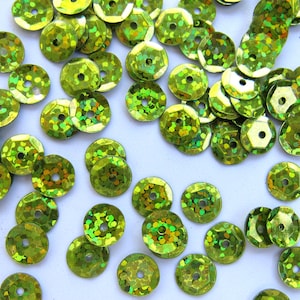 May include: Close-up of a collection of lime green sequins. The sequins are round with a central hole and a faceted surface, creating a sparkling effect. The sequins are scattered across a white surface.