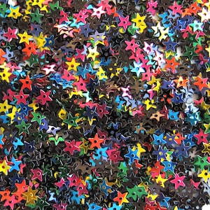 May include: A close-up shot of a collection of star-shaped sequins in various colors, including red, yellow, blue, green, and pink. The sequins have a metallic sheen and are scattered across the frame.