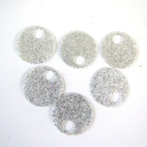 Op de afbeelding: Zes heldere, ronde bedels gevuld met zilveren glitter, elk met een klein gaatje aan de bovenkant. Deze sprankelende bedels zijn ideaal voor het maken van sieraden of knutselen. De bedels zijn in een cirkelvorm gerangschikt.