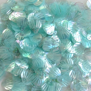 May include: A collection of iridescent, seashell-shaped sequins in aqua and pale blue. Each sequin has a scalloped edge and three small holes for sewing. The sequins are translucent, reflecting light. Ideal for craft projects.