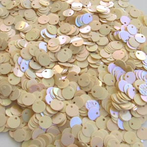 May include: A pile of iridescent, round sequins in a light beige color. Each sequin has a small hole near the top. The sequins have a subtle shimmer, reflecting hints of pink and blue. These craft supplies are ideal for embellishing clothing and accessories.