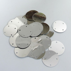 Könnte beinhalten: Eine Ansammlung kreisförmiger, silberfarbener Metallscheiben, die jeweils vier kleine, gleichmäßig verteilte Löcher nahe dem Rand aufweisen. Die Scheiben haben eine glatte, reflektierende, spiegelartige Oberfläche und liegen lose verstreut.
