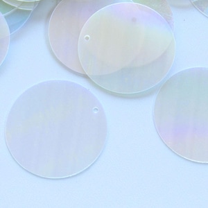 Peut inclure: Gros plan de plusieurs paillettes rondes et irisées. Chaque paillette a un petit trou près du bord. Les paillettes ont un effet subtil et chatoyant, reflétant une gamme de couleurs pastel. Elles sont dispersées sur une surface claire.