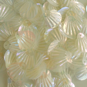 Pode incluir: Uma coleção de lantejoulas iridescentes em forma de concha. As lantejoulas são de uma cor pálida e translúcida com um brilho sutil, que capta a luz para criar um efeito perolado. Cada lantejoula tem um pequeno orifício para costurar ou enfiar.