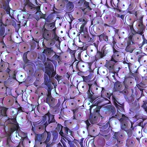 May include: A pile of iridescent purple sequins. Each sequin is round with a hexagonal hole in the center. The sequins have a shiny, reflective surface, creating a shimmering effect. The sequins are scattered, filling the frame.