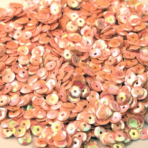 May include: A pile of iridescent peach-colored sequins. Each sequin is round with a central hole, and they vary slightly in color and sheen. The sequins are scattered, creating a textured, shimmering effect, suitable for crafts and embellishments.