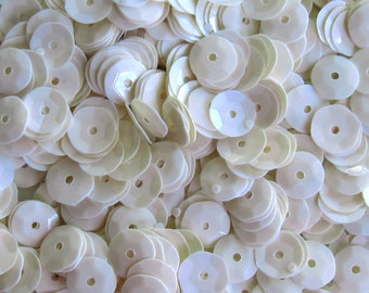 May include: A pile of ivory-colored sequins. Each sequin is round with a central hole and a faceted edge, creating a shimmering effect. The sequins are scattered, filling the frame and reflecting light.