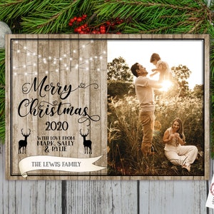May include: A wooden Christmas sign with the text "Merry Christmas 2020" and a family photo. The sign includes the family's name and deer silhouettes. Festive greenery and ornaments surround the sign.