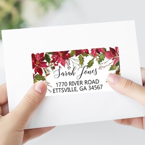 May include: A rectangular address label on a white envelope featuring a watercolor poinsettia and holly leaf border. The text reads Sarah Jones, 1770 River Road, Ettsville, GA 34567 in black script and sans-serif fonts.
