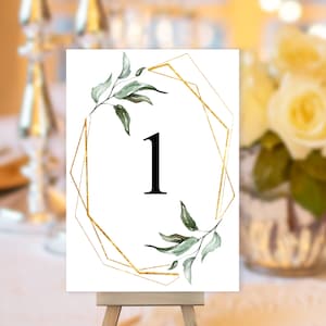 May include: White table number card with a gold geometric frame and green foliage. The number "1" is displayed in black font.