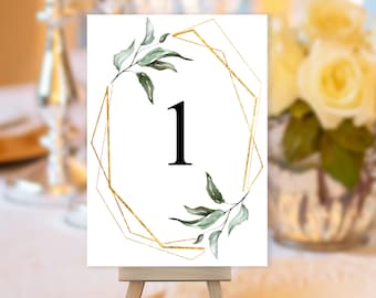 May include: White table number card with a gold geometric frame and green foliage. The number "1" is displayed in black font.