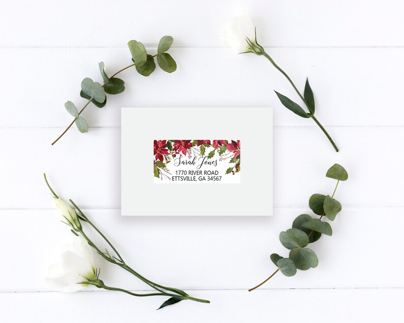 May include: A rectangular white mailing label featuring a festive red poinsettia and green holly leaf border. The label displays the printed text Sarah Jones, 1770 River Road, Ettsville, GA 34567 in a black serif and script font.