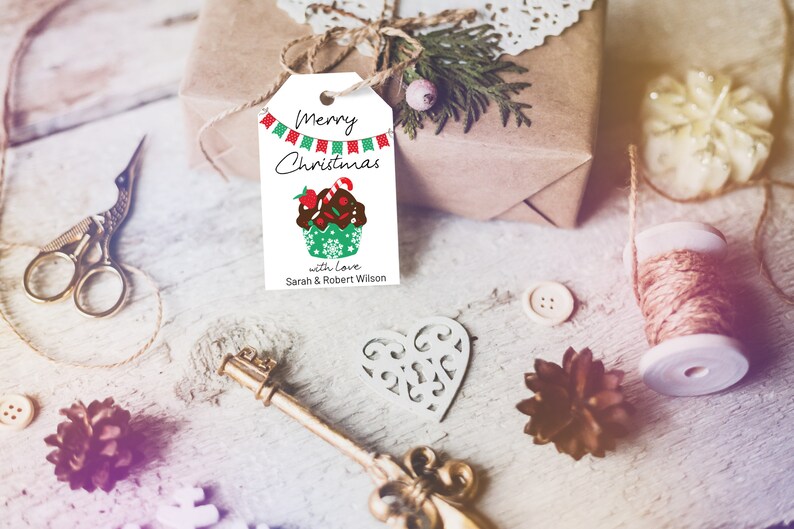 Pode incluir: Uma etiqueta de presente retangular branca com um furo no topo, apresentando uma guirlanda de bandeirinhas vermelhas e verdes, o texto Merry Christmas, um gr&aacute;fico de um cupcake de chocolate com uma bengala doce e o texto with love Sarah & Robert Wilson.
