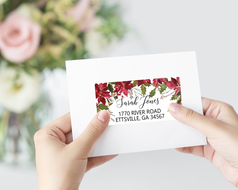 May include: A rectangular address label on a white envelope featuring a watercolor poinsettia and holly leaf border. The text reads Sarah Jones, 1770 River Road, Ettsville, GA 34567 in black script and sans-serif fonts.