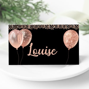 Pode incluir: Um cartão de lugar preto e ouro rosa com uma borda de confete ouro rosa. O cartão apresenta dois balões ouro rosa com brilho e o nome "Louise" escrito em ouro rosa.