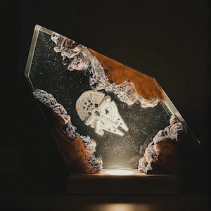 May include: A decorative lamp featuring a miniature white spaceship model suspended within a clear resin block. The resin is framed by jagged, natural brown wood edges, creating a space-like scene with embedded white speckles.