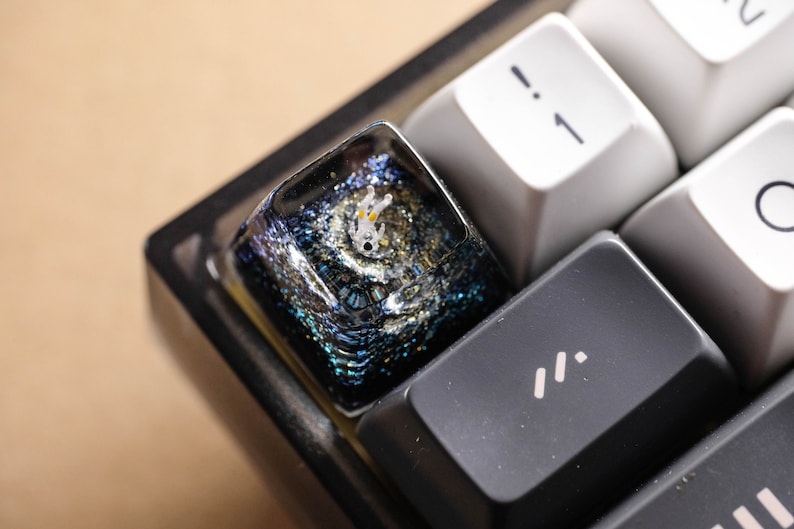 Peut inclure: Une touche de clavier m&eacute;canique personnalis&eacute;e avec une base en r&eacute;sine bleu fonc&eacute; et noire remplie de paillettes scintillantes. Une petite figurine d'astronaute blanche et translucide est incrust&eacute;e au centre, entour&eacute;e d'un effet spatial &eacute;toil&eacute;.