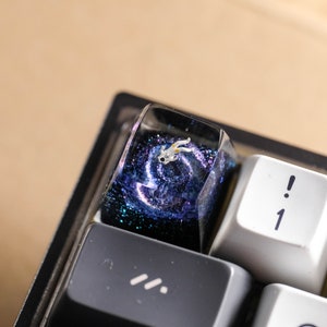 Peut inclure: Une touche de clavier artisanale personnalis&eacute;e avec un design en r&eacute;sine inspir&eacute; d'une galaxie bleu fonc&eacute; et violette avec des paillettes. Une petite figurine d'astronaute blanche d&eacute;taill&eacute;e est incrust&eacute;e dans la r&eacute;sine transparente.
