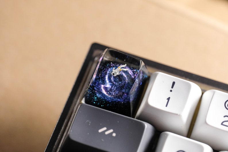 Peut inclure: Une touche de clavier artisanale personnalis&eacute;e avec un design en r&eacute;sine inspir&eacute; d'une galaxie bleu fonc&eacute; et violette avec des paillettes. Une petite figurine d'astronaute blanche d&eacute;taill&eacute;e est incrust&eacute;e dans la r&eacute;sine transparente.