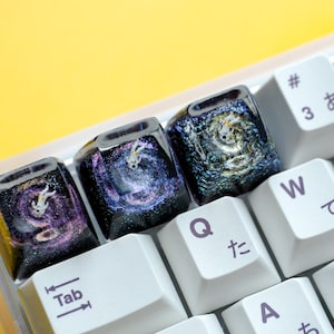 Peut inclure: Trois touches de clavier artisanales personnalis&eacute;es avec une base en r&eacute;sine noire profonde incrust&eacute;e de paillettes et une figurine d'astronaute blanche flottant dans un design de galaxie violet et bleu tourbillonnant.