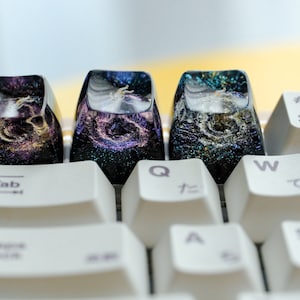 Peut inclure: Trois touches de clavier personnalis&eacute;es en r&eacute;sine avec un motif de galaxie spatiale, int&eacute;grant des paillettes violettes, dor&eacute;es et bleues dans une finition translucide sombre, install&eacute;es sur un clavier m&eacute;canique blanc avec des touches standard.