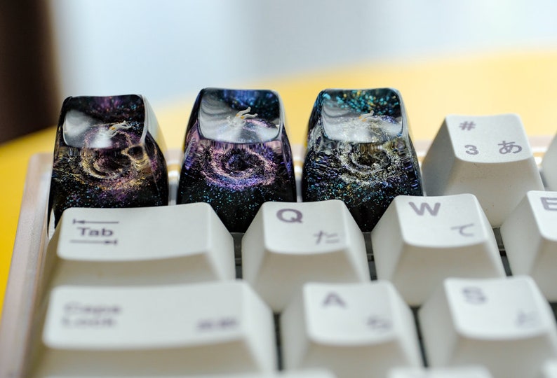 Peut inclure: Trois touches de clavier personnalis&eacute;es en r&eacute;sine avec un motif de galaxie spatiale, int&eacute;grant des paillettes violettes, dor&eacute;es et bleues dans une finition translucide sombre, install&eacute;es sur un clavier m&eacute;canique blanc avec des touches standard.