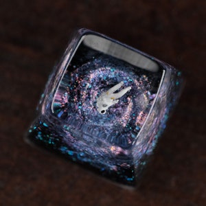 Peut inclure: Une touche de clavier carr&eacute;e en r&eacute;sine translucide contenant une figurine d'astronaute blanche miniature suspendue dans un design de galaxie sombre et tourbillonnant. L'int&eacute;rieur pr&eacute;sente des paillettes iris&eacute;es aux reflets bleus et violets.