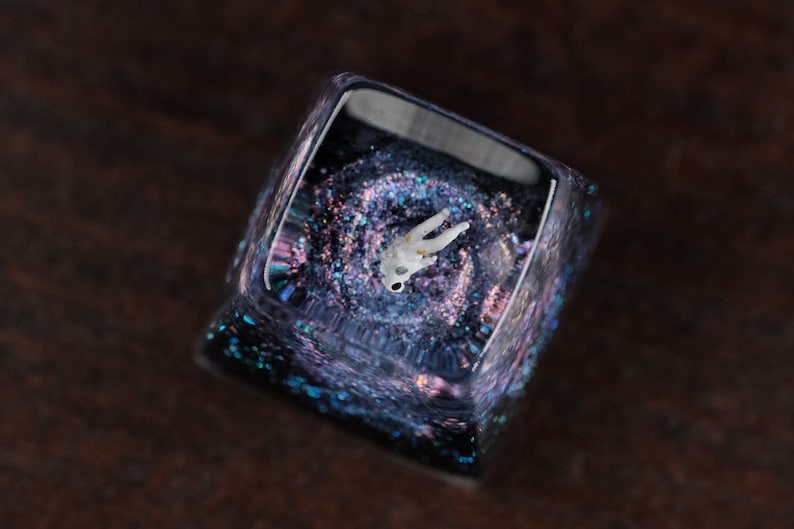 Peut inclure: Une touche de clavier carr&eacute;e en r&eacute;sine translucide contenant une figurine d'astronaute blanche miniature suspendue dans un design de galaxie sombre et tourbillonnant. L'int&eacute;rieur pr&eacute;sente des paillettes iris&eacute;es aux reflets bleus et violets.