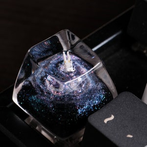 Peut inclure: Une touche de clavier m&eacute;canique en r&eacute;sine transparente avec un int&eacute;rieur rempli de paillettes bleu fonc&eacute; et violet &eacute;voquant une n&eacute;buleuse. Une petite figurine blanche avec des accents orange est incrust&eacute;e au centre.