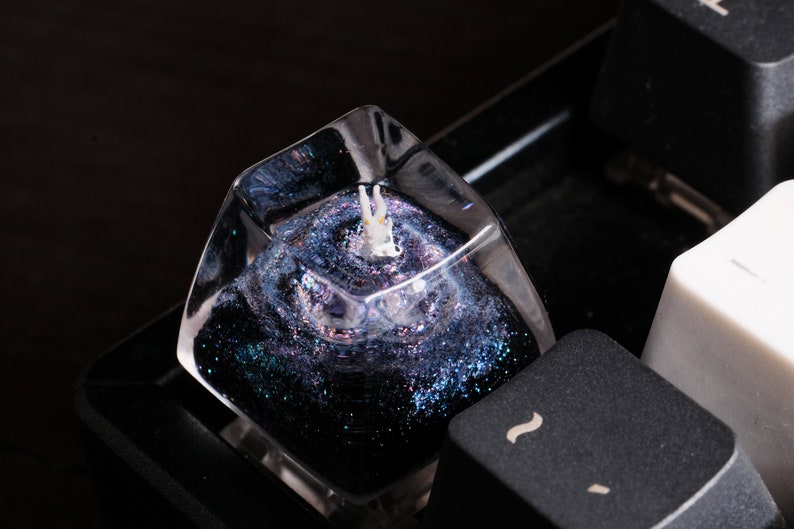 Peut inclure: Une touche de clavier m&eacute;canique en r&eacute;sine transparente avec un int&eacute;rieur rempli de paillettes bleu fonc&eacute; et violet &eacute;voquant une n&eacute;buleuse. Une petite figurine blanche avec des accents orange est incrust&eacute;e au centre.