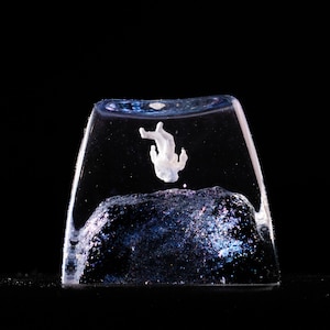 Peut inclure: Une touche de clavier m&eacute;canique en r&eacute;sine transparente contenant une petite figurine d'astronaute blanche en suspension au-dessus d'une base sombre et scintillante aux reflets bleu fonc&eacute; et violets, sur un fond noir uni.