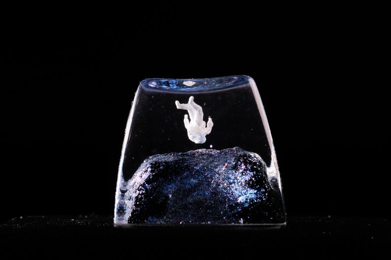 Peut inclure: Une touche de clavier m&eacute;canique en r&eacute;sine transparente contenant une petite figurine d'astronaute blanche en suspension au-dessus d'une base sombre et scintillante aux reflets bleu fonc&eacute; et violets, sur un fond noir uni.