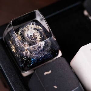 Peut inclure: Une touche de clavier m&eacute;canique en r&eacute;sine transparente pr&eacute;sentant un motif d'espace profond avec des tourbillons bleu fonc&eacute; et noirs, des paillettes dor&eacute;es et une petite figurine blanche &agrave; deux cornes incrust&eacute;e dans le mat&eacute;riau.