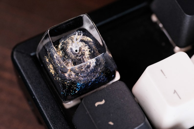 Peut inclure: Une touche de clavier m&eacute;canique en r&eacute;sine transparente pr&eacute;sentant un motif d'espace profond avec des tourbillons bleu fonc&eacute; et noirs, des paillettes dor&eacute;es et une petite figurine blanche &agrave; deux cornes incrust&eacute;e dans le mat&eacute;riau.