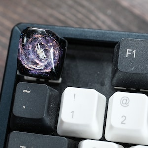 Peut inclure: Une touche de clavier m&eacute;canique en r&eacute;sine personnalis&eacute;e repr&eacute;sentant une figurine d'astronaute blanche flottant dans un design de galaxie tourbillonnant violet fonc&eacute; et noir avec des paillettes, plac&eacute;e sur un clavier noir pr&egrave;s des touches F1.