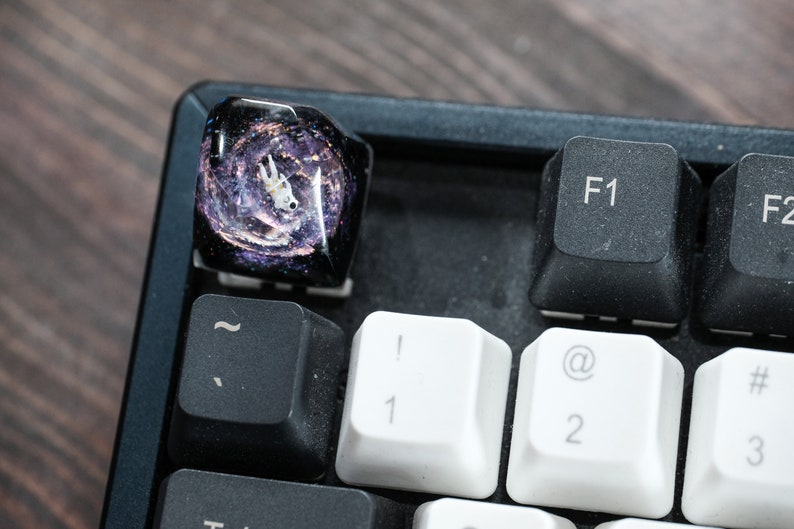 Peut inclure: Une touche de clavier m&eacute;canique en r&eacute;sine personnalis&eacute;e repr&eacute;sentant une figurine d'astronaute blanche flottant dans un design de galaxie tourbillonnant violet fonc&eacute; et noir avec des paillettes, plac&eacute;e sur un clavier noir pr&egrave;s des touches F1.