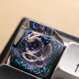 Peut inclure: Une touche de clavier m&eacute;canique en r&eacute;sine transparente contenant une figurine d'astronaute blanc miniature suspendue dans un d&eacute;cor spatial bleu et violet rempli de paillettes, plac&eacute;e sur un clavier noir &agrave; c&ocirc;t&eacute; d'une touche blanche.