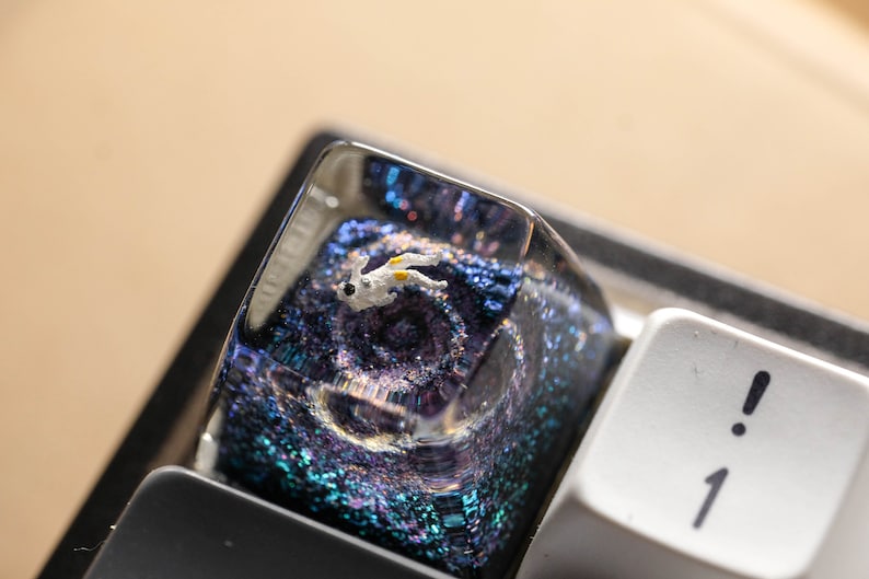 Peut inclure: Une touche de clavier m&eacute;canique en r&eacute;sine transparente contenant une figurine d'astronaute blanc miniature suspendue dans un d&eacute;cor spatial bleu et violet rempli de paillettes, plac&eacute;e sur un clavier noir &agrave; c&ocirc;t&eacute; d'une touche blanche.