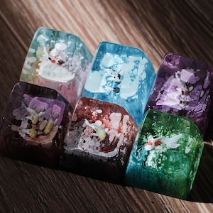 Peut inclure: Un ensemble de six touches de clavier mécanique en résine translucide, chacune présentant un poisson koï miniature nageant dans un environnement aquatique texturé et coloré. Les touches arborent des teintes bleues, violettes, vertes et roses.