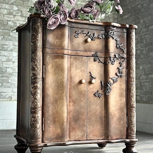 May include: A vintage-style wooden cabinet with a distressed bronze finish. The cabinet features ornate details, including floral accents and bird motifs. It has two doors and a drawer, with decorative hardware. Artificial purple roses adorn the top. The cabinet is a piece of furniture.