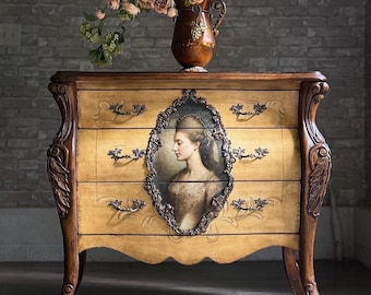 May include: A wooden chest of drawers with a light yellow finish, featuring three drawers with dark metal handles and a central oval portrait of a person in profile. The piece has curved legs and ornate carvings. A copper-colored vase with flowers sits on top.