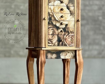May include: A wooden jewelry armoire with a floral rose design on the drawer fronts. The armoire has a gold-colored trim and sits on four curved legs. The top of the armoire has a decorative floral accent.