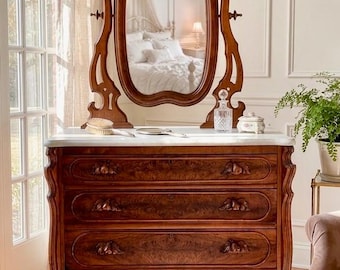May include: A wooden dresser with four drawers featuring carved handles and a white marble top. A matching oval swivel mirror with an ornate wooden frame sits on the surface. The wood has a rich, dark brown grain pattern throughout the structure.