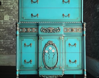 May include: A teal blue dresser with gold accents and floral details. The dresser has six drawers and sits on four legs. The dresser is labeled "ReLove ReStore".