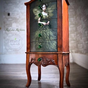 May include: A wooden jewelry armoire with five drawers and green glass knobs. The front of the armoire is decorated with a print of a fairy with green wings and a green dress. The armoire is standing on four legs.