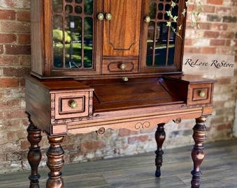 May include: A dark wood secretary desk featuring a hutch with two glass-paned doors, a central cabinet, and a small drawer. The base includes a drop-down writing surface, two side drawers, and four turned legs. The text ReLove ReStore is visible.
