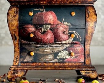 May include: A three-drawer chest with a dark, distressed gold finish and a graphic print of red apples in a bowl across the drawer fronts. The chest features small, round, light-colored wooden knobs and the text ReLove ReStore.