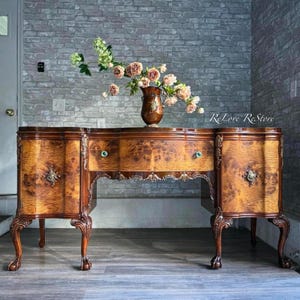 May include: A vintage wooden sideboard with intricate carvings and a burlwood veneer. It has three drawers and two doors with brass hardware. The sideboard is sitting on a wood floor in front of a brick wall. A vase with pink flowers is on top of the sideboard.